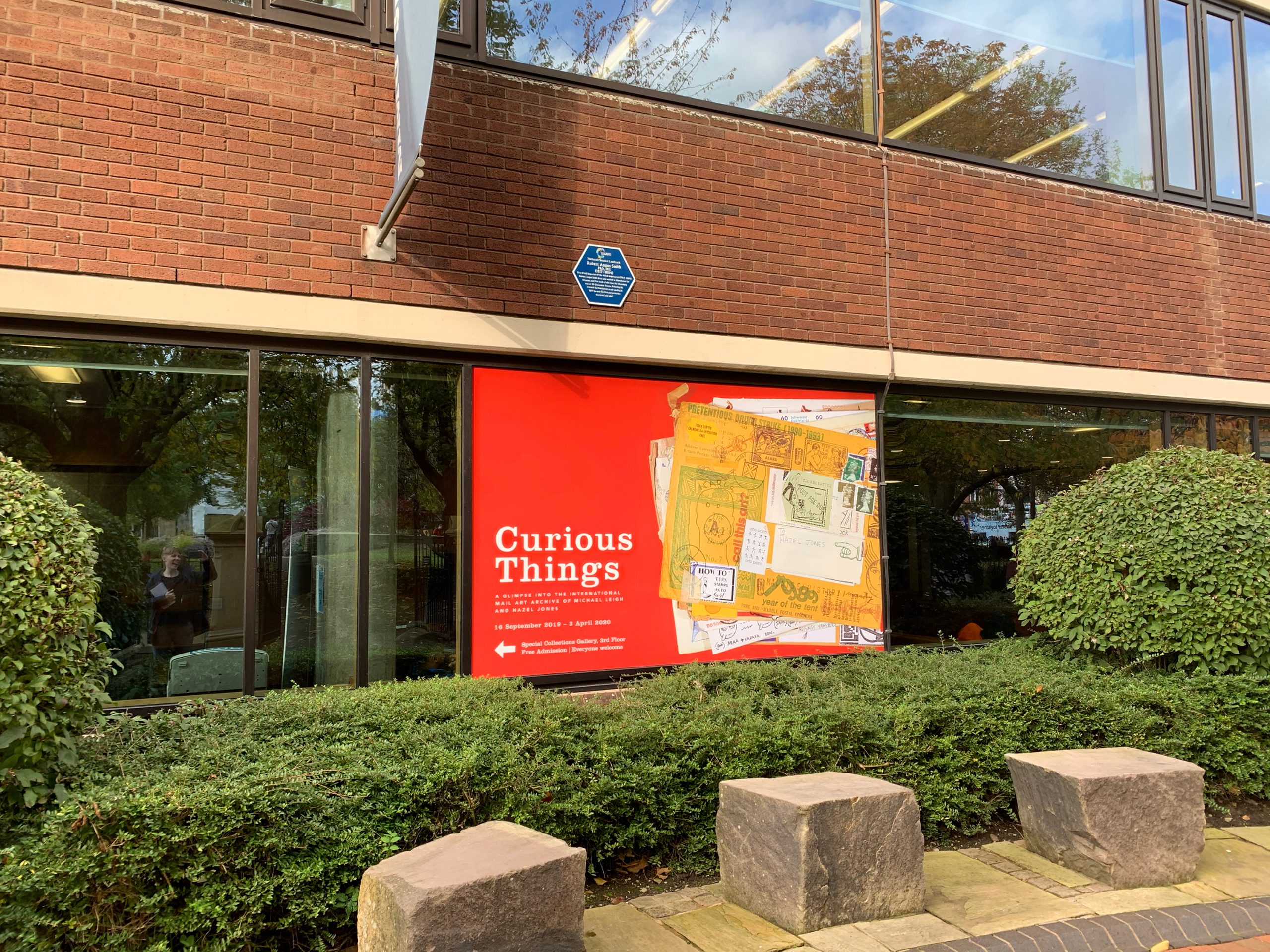 front of library building surrounded by bushes with red window vinyl with stack of mail art envelopes advertising Curious Things exhibition